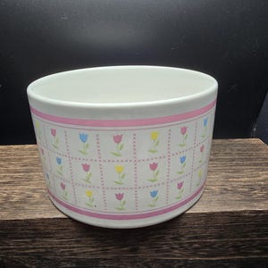May include: A white ceramic bowl with a pink band and a repeating pattern of colorful tulips. The tulips are pink, yellow, and blue, set against a grid of small crosses. The bowl sits on a dark wooden surface.