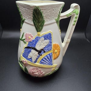 May include: Cream-colored ceramic pitcher with a decorative dragonfly and floral design. The pitcher features a blue and yellow fan, pink and orange flowers, and green leaves. The handle is adorned with green leaves.