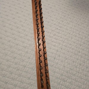 May include: A long, rectangular wooden board featuring two parallel rows of evenly spaced metal pegs. The wood has a visible grain pattern in a medium brown tone, and the metal pegs are dark in color with rounded, knob-like tops.