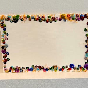 May include: A white card with a colorful border of sequins. The sequins are arranged in a frame around the edge of the card.