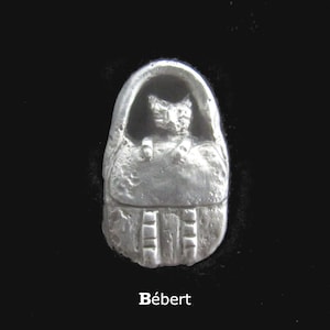 May include: A silver-colored pendant featuring a cat inside a hooded carrier. The pendant has a textured surface and the word "Bébert" is printed at the bottom. The background is black.