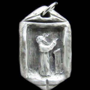May include: A silver pendant with a raised image of a person in a robe holding a book. The pendant is in the shape of a rectangle with rounded corners.