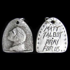 May include: Two silver pendants, one with a profile of a person's face and the other with the text "MATT TALBOT PRAY FOR US" engraved on it.