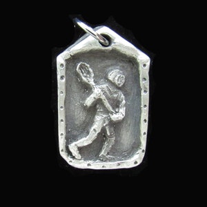 May include: Silver charm featuring a lacrosse player in action, with a detailed design and a textured border.