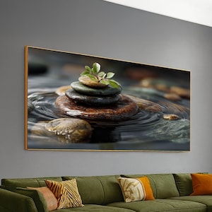 May include: A framed canvas print featuring a stack of smooth, flat stones in water, topped with a small green plant. The stones are various shades of brown and gray, creating a zen-like composition. The water has ripples.
