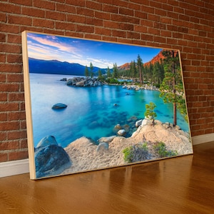 Puede incluir: Impresión en lienzo enmarcada de un paisaje lacustre escénico. La imagen presenta agua turquesa clara, orillas rocosas y árboles verdes. Montañas y un cielo azul con nubes son visibles en el fondo. El marco es de color madera clara.