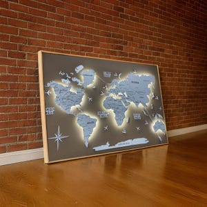 Pode incluir: Um mapa-múndi emoldurado com fundo cinza e continentes azuis. O mapa é iluminado com luzes LED, destacando os continentes e oceanos. Ícones decorativos de avião e bússola estão incluídos. A moldura é de madeira clara.