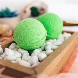 May include: Two bright green bath bombs rest on white pebbles inside a small wooden tray. The tray sits on a pink and orange fabric. A small succulent and other decorative items are in the background.