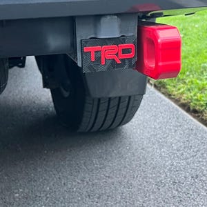 May include: A black hitch cover featuring a textured diamond plate pattern and the red TRD logo in a bold font. The cover is mounted on a vehicle receiver hitch next to a bright red metal tow hook, positioned above a black tyre and wheel assembly.