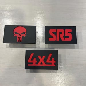 May include: Three black rectangular emblems with red designs. One features a skull, another reads "SR5", and the third says "4X4". The emblems are likely for automotive customization.