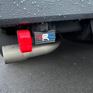 May include: A rectangular hitch cover featuring a grey American flag pattern with the letters TRD in red, white, and blue. The cover is mounted on a vehicle bumper next to a bright red tow hook and a metallic exhaust pipe, all covered in raindrops.