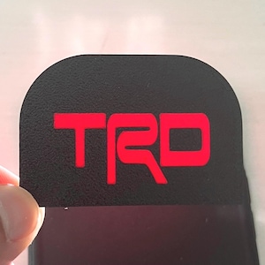 以下が含まれることがあります： 赤い「TRD」の文字が特徴的な、上部が丸みを帯びた黒い長方形のエンブレム。背景はぼやけており、手で持たれています。文字は太字で、黒い背景に映えます。