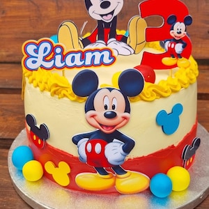 May include: A Mickey Mouse themed birthday cake. The cake features a yellow base with red and yellow detailing, and blue and yellow sweets. The name "Liam" is written on the cake, along with the number "3".