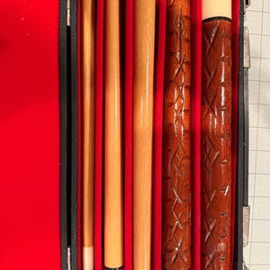 May include: A pool cue set in a black case with a red velvet interior. The set includes multiple wooden cue sticks, some with intricate carved designs. The cues are various shades of brown and tan, with white tips. The case is open, displaying the cues.
