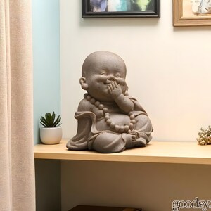 K&ouml;nnte beinhalten: Eine steinfarbene Buddha-Statue in sitzender Position, mit einer Hand, die den Mund bedeckt, als w&uuml;rde sie lachen. Die Statue hat eine Perlenkette und steht auf einem Holzregal. Links ist eine kleine Topfpflanze.