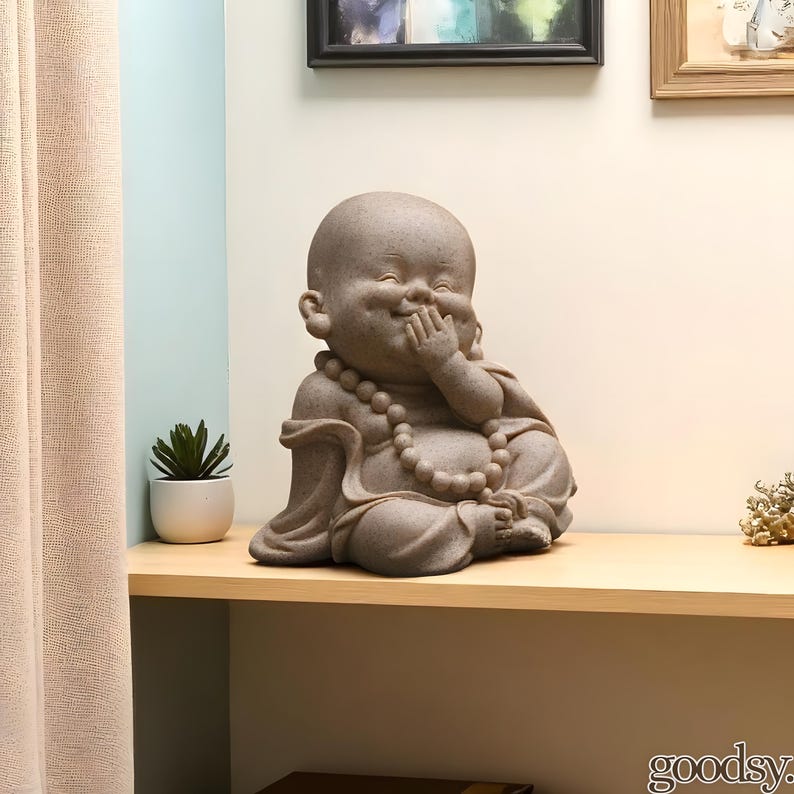 K&ouml;nnte beinhalten: Eine steinfarbene Buddha-Statue in sitzender Position, mit einer Hand, die den Mund bedeckt, als w&uuml;rde sie lachen. Die Statue hat eine Perlenkette und steht auf einem Holzregal. Links ist eine kleine Topfpflanze.