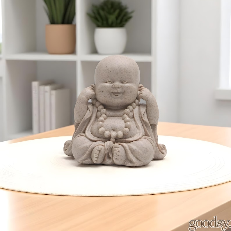 K&ouml;nnte beinhalten: Kleine, graue Buddha-Statue in sitzender Position, mit den H&auml;nden an den Ohren und einem l&auml;chelnden Gesichtsausdruck. Die Statue hat eine Perlenkette und steht auf einer wei&szlig;en, kreisf&ouml;rmigen Oberfl&auml;che. Der Hintergrund umfasst einen Holztisch, ein wei&szlig;es Regal und Topfpflanzen.