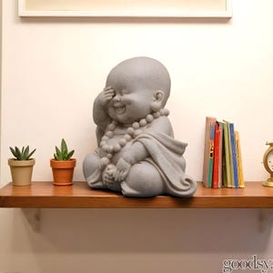 K&ouml;nnte beinhalten: Eine graue Stein-Buddha-Statue mit l&auml;chelndem Gesichtsausdruck, die ein Auge mit einer Hand bedeckt. Die Statue ist mit einer Perlenkette geschm&uuml;ckt und in ein Gewand geh&uuml;llt. Zwei kleine Topfpflanzen und ein Stapel B&uuml;cher stehen auf einem Holzregal.