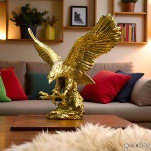 Peut inclure: Une sculpture d'aigle doré aux ailes déployées, perchée sur une base texturée. La statue d'oiseau détaillée est un élément de décoration intérieure, avec une finition métallique. L'aigle est exposé sur une table en bois devant un canapé avec des coussins colorés.