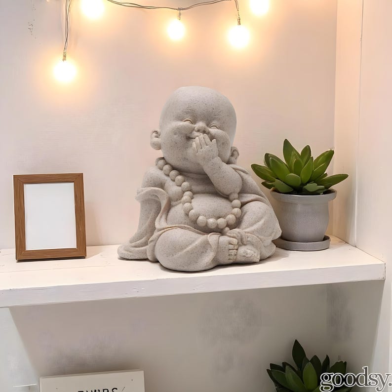 K&ouml;nnte beinhalten: Eine hellgraue Buddha-Statue mit einer Perlenkette, die mit einer Hand vor dem Mund sitzt. Ein kleiner Holzbilderrahmen und eine Topf-Sukkulente befinden sich ebenfalls auf dem Regal. Dekorative Lichterketten befinden sich dar&uuml;ber.