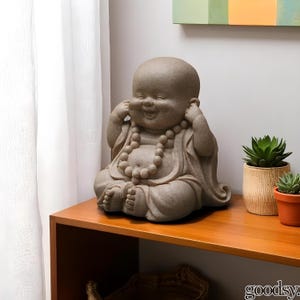 K&ouml;nnte beinhalten: Eine l&auml;chelnde, sitzende Buddha-Statue mit den H&auml;nden an den Ohren. Die Statue ist hellbeige und tr&auml;gt eine Perlenkette. Sie steht auf einem Holzregal neben Topf-Sukkulenten. Ein dekorativer Artikel.