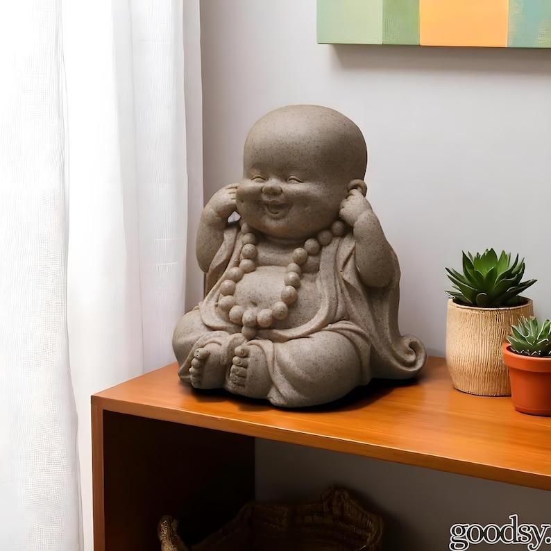 K&ouml;nnte beinhalten: Eine l&auml;chelnde, sitzende Buddha-Statue mit den H&auml;nden an den Ohren. Die Statue ist hellbeige und tr&auml;gt eine Perlenkette. Sie steht auf einem Holzregal neben Topf-Sukkulenten. Ein dekorativer Artikel.