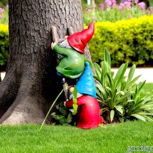 May include: A garden gnome statue depicting three gnomes ascending a wooden ladder. The gnomes are dressed in red, green, and blue outfits. The statue is positioned near a tree trunk, set against a backdrop of green grass and plants.