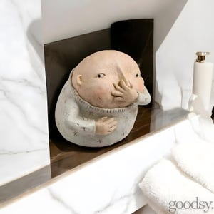 May include: A ceramic figurine of a person with a large head and a hand covering their nose. The figurine is wearing a gray sweater with a white star pattern.