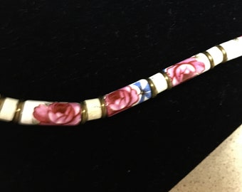 May include: A close-up of a necklace featuring white, rectangular beads adorned with pink rose floral patterns and blue accents. The beads are separated by gold-colored metal rings, creating a delicate and ornate design. The necklace is set against a dark background.