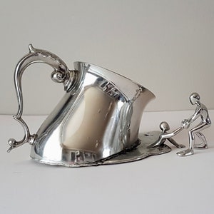 Könnte beinhalten: Eine silberne Teekanne mit einem Vogelgriff, die schmilzt und tropft, mit zwei silbernen Figuren, die helfen, sie anzuheben.