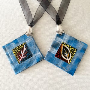 May include: Two handmade square pendants with blue and white plaid backgrounds. Each pendant has a smaller square with a leaf design in red, white, and yellow. The pendants are attached to black ribbon necklaces.