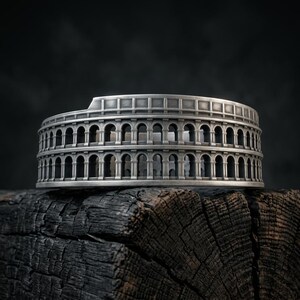 May include: A silver-toned ring featuring a detailed architectural design inspired by the Roman Colosseum. The band displays two tiers of repeating arched columns and a flat top edge, crafted with a weathered, metallic finish throughout the piece.