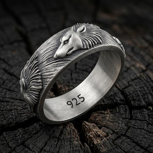 May include: A silver-toned ring featuring a relief design of wolf heads with detailed fur textures. The interior of the band is engraved with the number 925, indicating the metal purity. The surface of the ring has a brushed, matte finish.