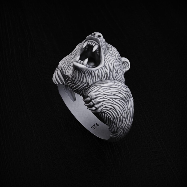 May include: A silver ring featuring a detailed, roaring bear head. The bear's mouth is open, showing sharp teeth. The ring is marked with the number "925" on the band.
