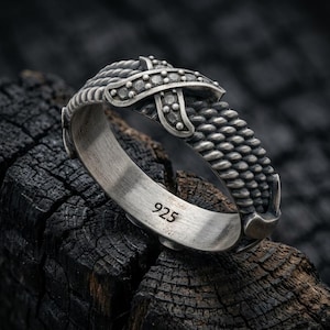 May include: A sterling silver ring featuring a textured, rope-like band with a prominent, raised cross-shaped overlay adorned with small, dark, circular indentations. The interior of the band is smooth and clearly stamped with the number 925.