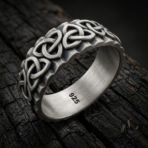 May include: A silver-toned ring featuring a continuous embossed triquetra knot pattern around the outer band. The interior of the ring has a brushed metal finish and is clearly stamped with the number 925, indicating its sterling silver composition.