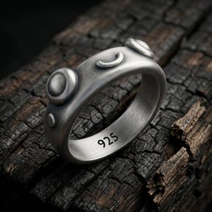 Könnte beinhalten: Ein silberner Ring mit gebürsteter Oberfläche, der erhabene kreisförmige und halbmondförmige Details auf dem äußeren Band aufweist. Die Innenseite des Rings ist deutlich mit der Zahl 925 gestempelt, was den Silbergehalt des Metalls angibt.