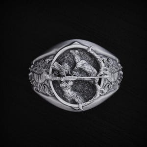 May include: A silver ring with a detailed design of a woman holding a bow and arrow, with a deer and other wildlife surrounding her.