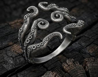 May include: An open-style ring crafted from dark, oxidized silver, featuring a detailed design of multiple octopus tentacles that wrap around the finger. The surface of the tentacles displays realistic suction cups and a textured, weathered finish.