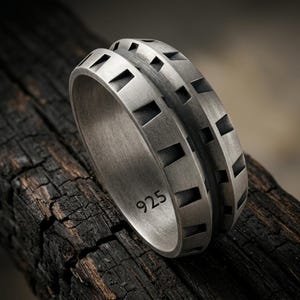 Puede incluir: Un anillo de plata cepillada con un borde exterior texturizado similar a un engranaje, con muescas trapezoidales repetitivas. El interior de la banda es liso y está claramente marcado con el número 925, indicando su composición de plata esterlina.