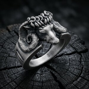 May include: A silver-toned ring featuring a detailed ram head sculpture with prominent curled horns. The metal surface has a matte, oxidized finish, and the inner band is clearly stamped with the number 925, indicating sterling silver composition.