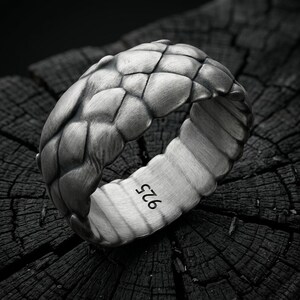 Puede incluir: Un anillo de plata con un patrón de escamas superpuestas y texturizadas en la superficie exterior. El interior de la banda es liso y está claramente marcado con el sello 925, indicando plata de ley. El acabado es de aspecto mate.