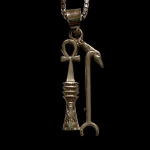 May include: Silver-toned pendant necklace featuring three Egyptian symbols: an Ankh, a Djed pillar, and a staff with a jackal head. The pendant hangs from a delicate chain against a black background.