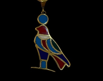 Puede incluir: Collar dorado con un colgante que presenta un diseño de pájaro estilizado y colorido. El pájaro tiene detalles en rojo, azul, verde y blanco, con un círculo azul sobre su cabeza. La cadena es de estilo eslabón delicado, sobre fondo negro.