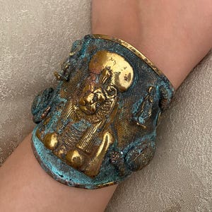 Könnte beinhalten: Ein breites Armband im antiken Stil mit detailliertem ägyptischem Design. Das Armband zeigt eine zentrale Figur mit Kopfschmuck, in Gold- und Bronzetönen, mit türkisfarbenen Patina-Akzenten. Das Armband wird am Handgelenk getragen.