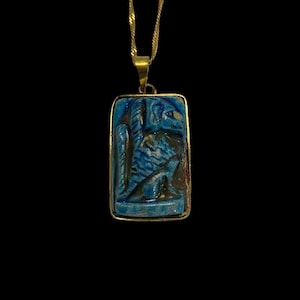 May include: A gold-colored chain necklace with a rectangular pendant. The pendant features a blue carved design with an ancient Egyptian theme. The pendant is framed in gold and hangs against a black background.