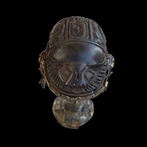 Könnte beinhalten: Eine dunkelgraue, aufwendig geschnitzte Skarabäus-Käfer-Skulptur mit Hieroglyphen auf dem Rücken. Der Käfer hat goldfarbene Akzente und ist auf einem silberfarbenen Sockel montiert. Der Hintergrund ist schwarz.
