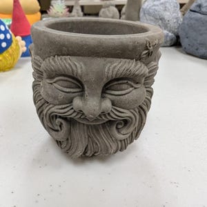 May include: A grey concrete planter shaped like a man's face with a beard. The planter has a wide, round opening at the top.