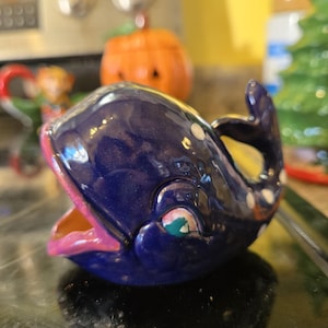 Peut inclure: Figurine de baleine en céramique bleu foncé, avec une bouche rose et des accents blancs et orange. La baleine a un design fantaisiste, avec un petit œil et une queue incurvée. Objet décoratif.