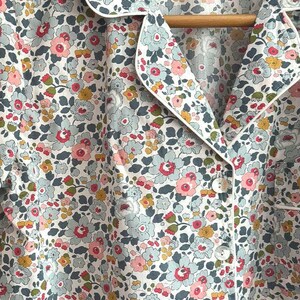Può includere: Top pigiama con stampa floreale con colletto e finiture bianche. Il tessuto presenta un motivo di piccoli fiori rosa, blu, gialli e verdi su sfondo bianco. La parte superiore ha un'abbottonatura e pende da un'appendiabiti di legno.